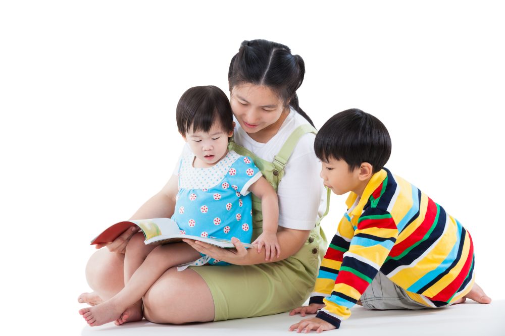 Image of cute young female with two little asian children reading a book together, daughter sitting on the lap, son sitting on the floor, happy family concept, isolated on white background